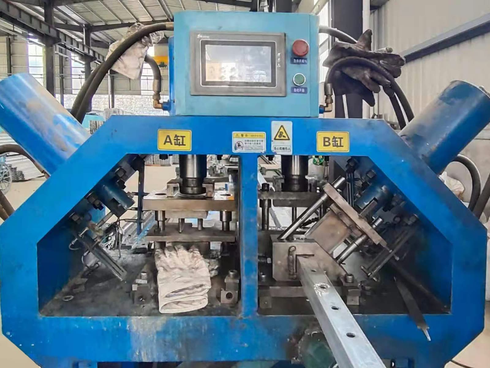 A blue hydraulic punching machine with a touch-screen control panel and two angled cylinders, punching holes in a square tube.