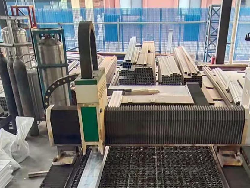 A flat-bed laser cutting machine with its gantry over a slatted cutting bed, gas cylinders at the left and stacks of square steel tubes behind.