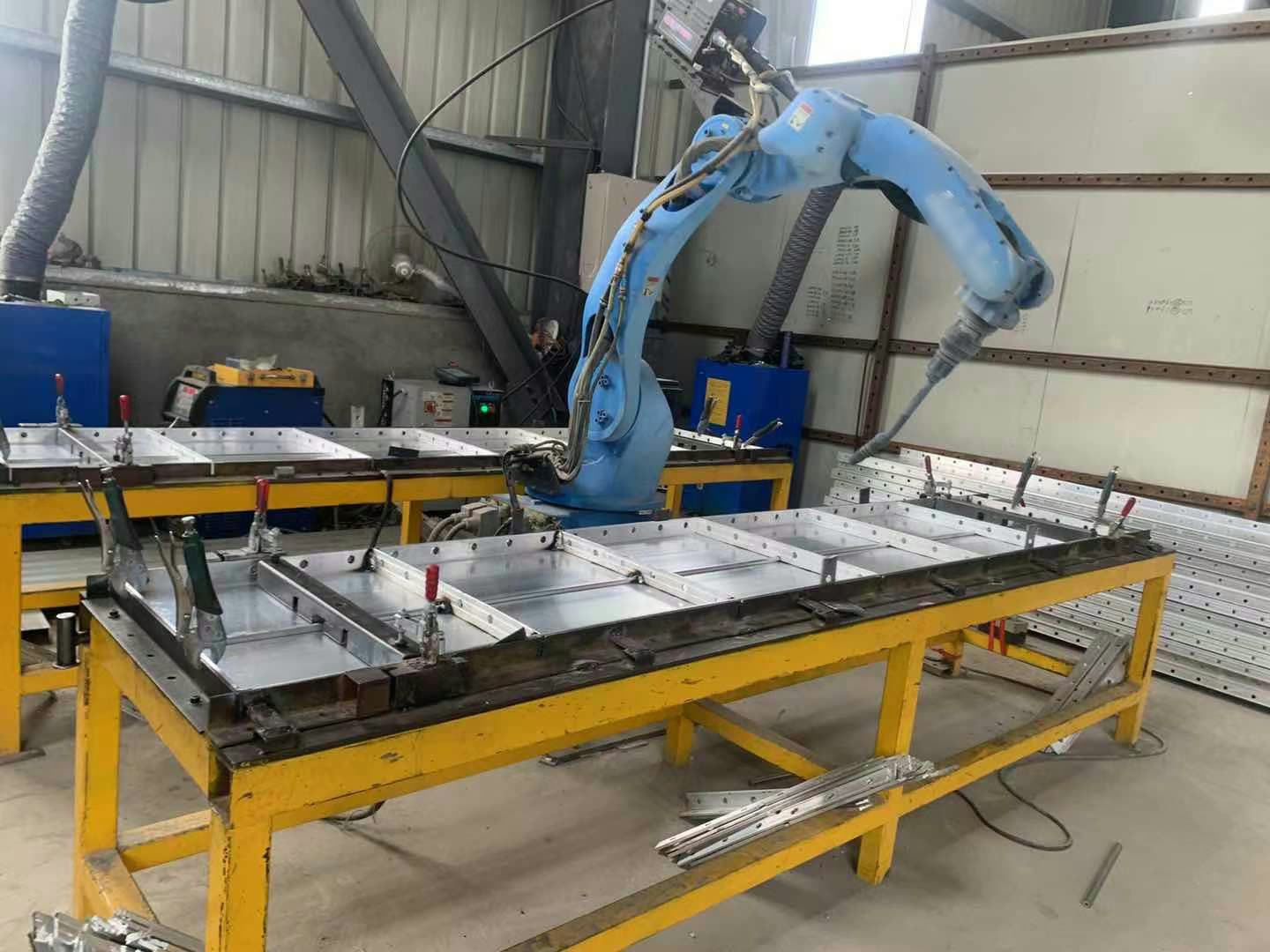 A blue industrial robot with a welding torch beside yellow fixture tables holding silver rectangular frames in clamps.