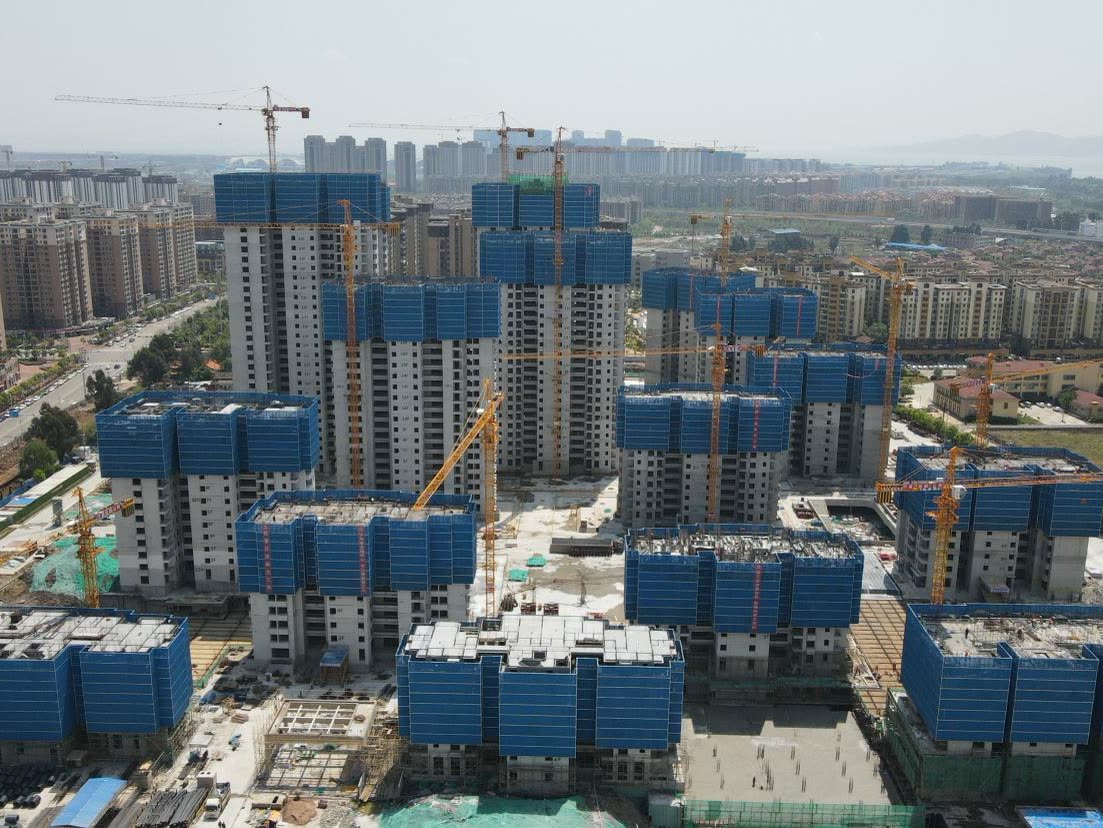 Aerial view of a residential development under construction: about a dozen high-rise towers with blue climbing-scaffold enclosures around their top floors, tower cranes between them and the city beyond.
