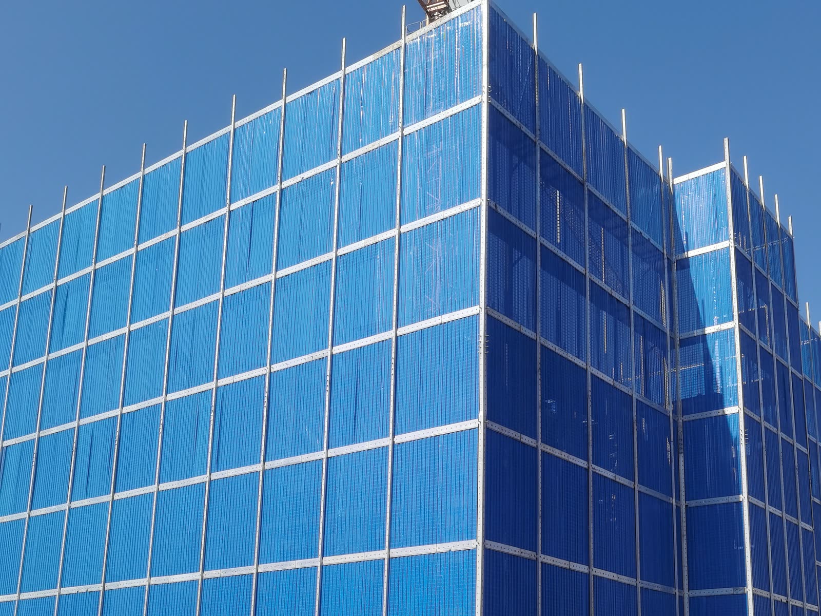 Corner of a tall blue mesh climbing-scaffold enclosure on a building under construction, with silver frame members rising above the panels against a blue sky.