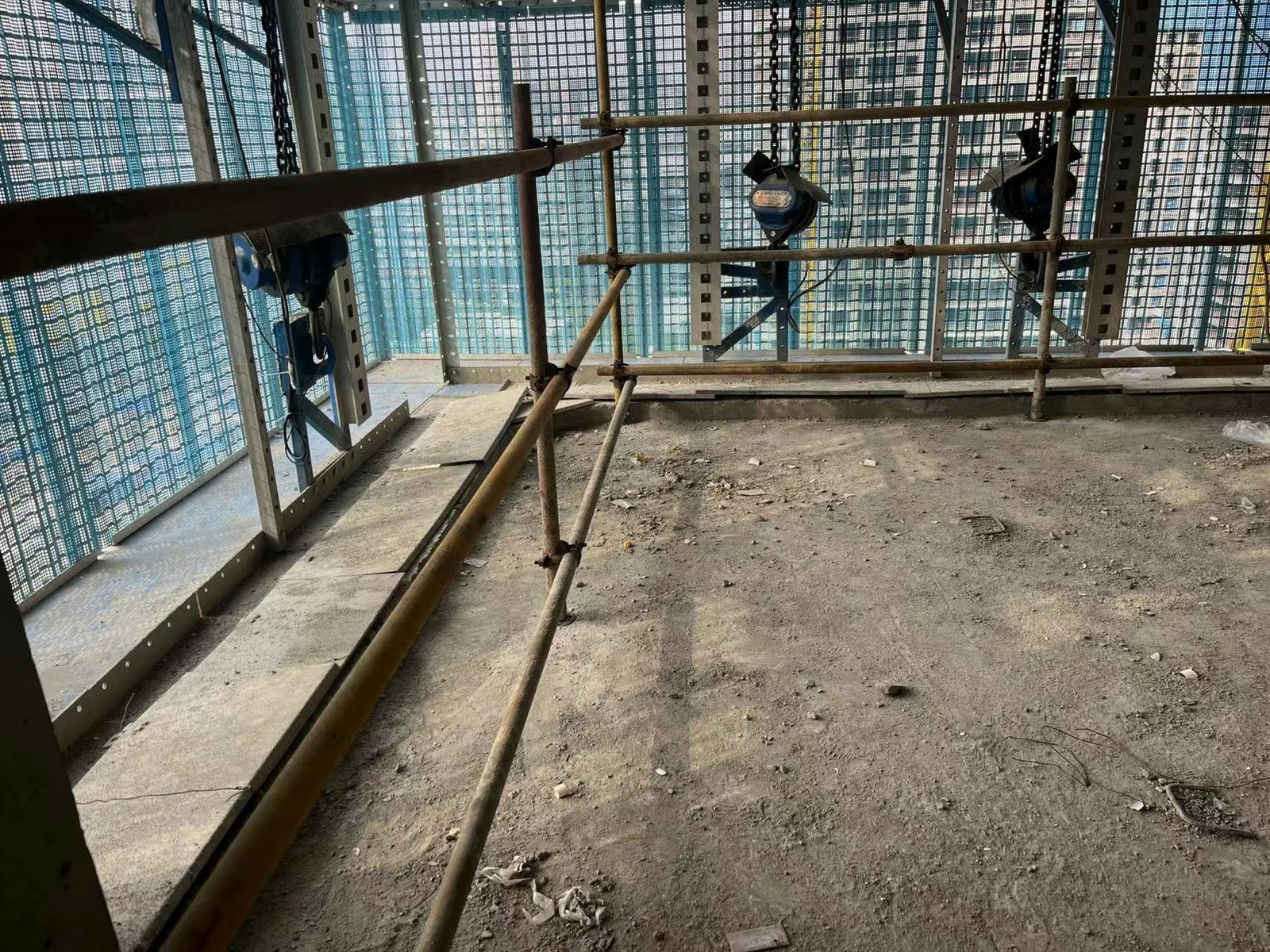 Seen from a bare floor slab: electric chain hoists hanging at the lifting points on slotted guide rails behind square-mesh perimeter panels, with temporary steel-tube guard rails in front.