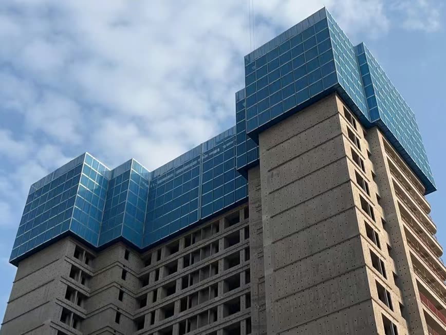 A high-rise residential tower under construction, its top storeys and stepped upper façade wrapped in a blue climbing-scaffold enclosure, against a cloudy sky.