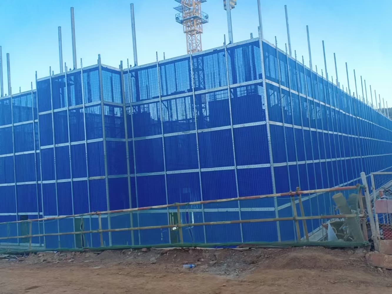 A blue mesh climbing-scaffold enclosure with silver frame members rising above it, assembled around the lower floors of a building at the start of construction, with a tower crane behind.