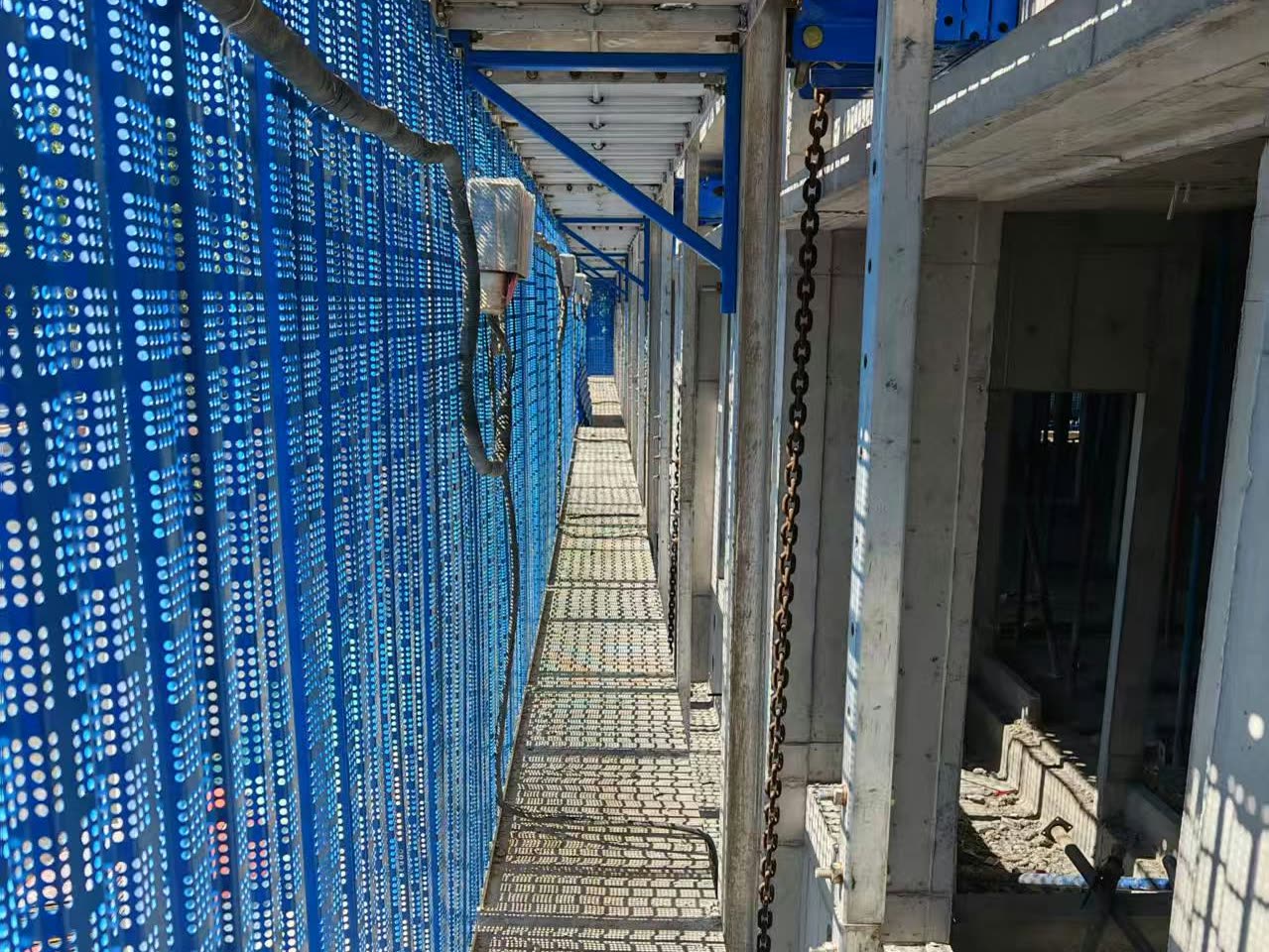 Inside a climbing scaffold: a perforated walkway deck runs between blue perforated mesh panels and the concrete building, with a hoist chain, cables and a blue support bracket overhead.
