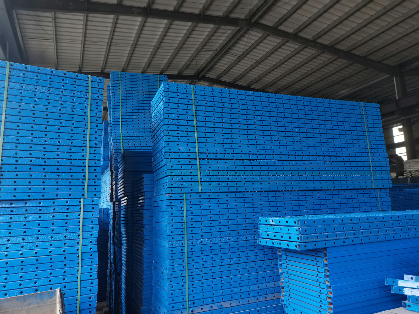 Tall strapped stacks of blue-painted perforated steel members under the roof of a portal-frame warehouse.