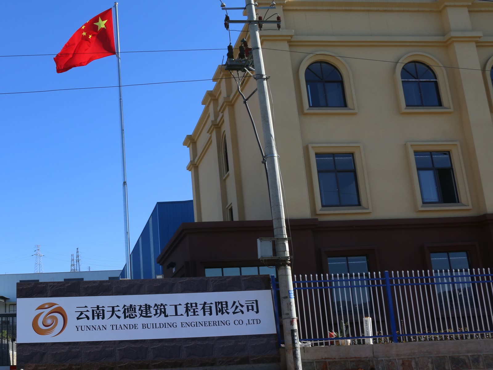 Entrance to TianDe’s premises: a sign with the company emblem and its Chinese and English names, a flagpole, a three-storey office building and a blue steel-clad workshop behind.