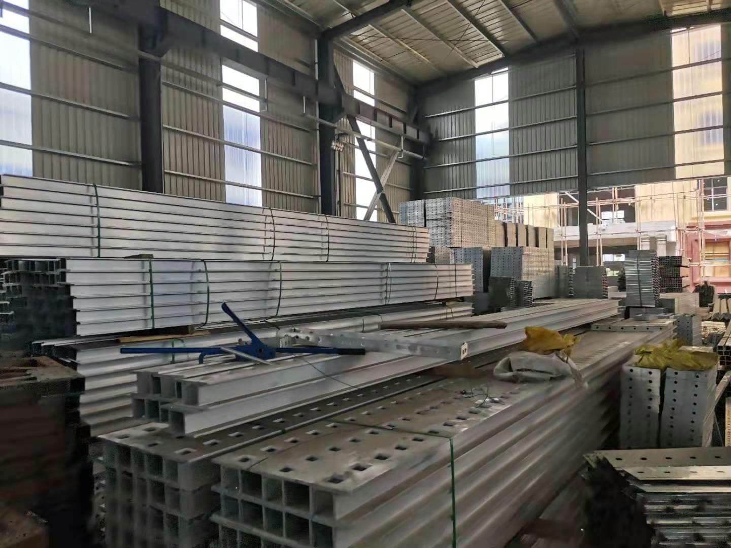 Strapped bundles of long aluminium-alloy extrusions and stacks of perforated metal components inside a portal-frame workshop.