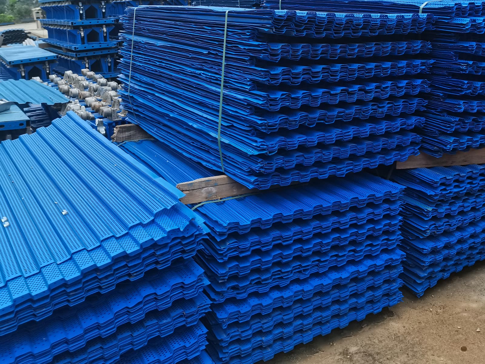 Stacks of blue perforated, corrugated mesh panels in an open yard, with blue wall-support assemblies stacked behind them.