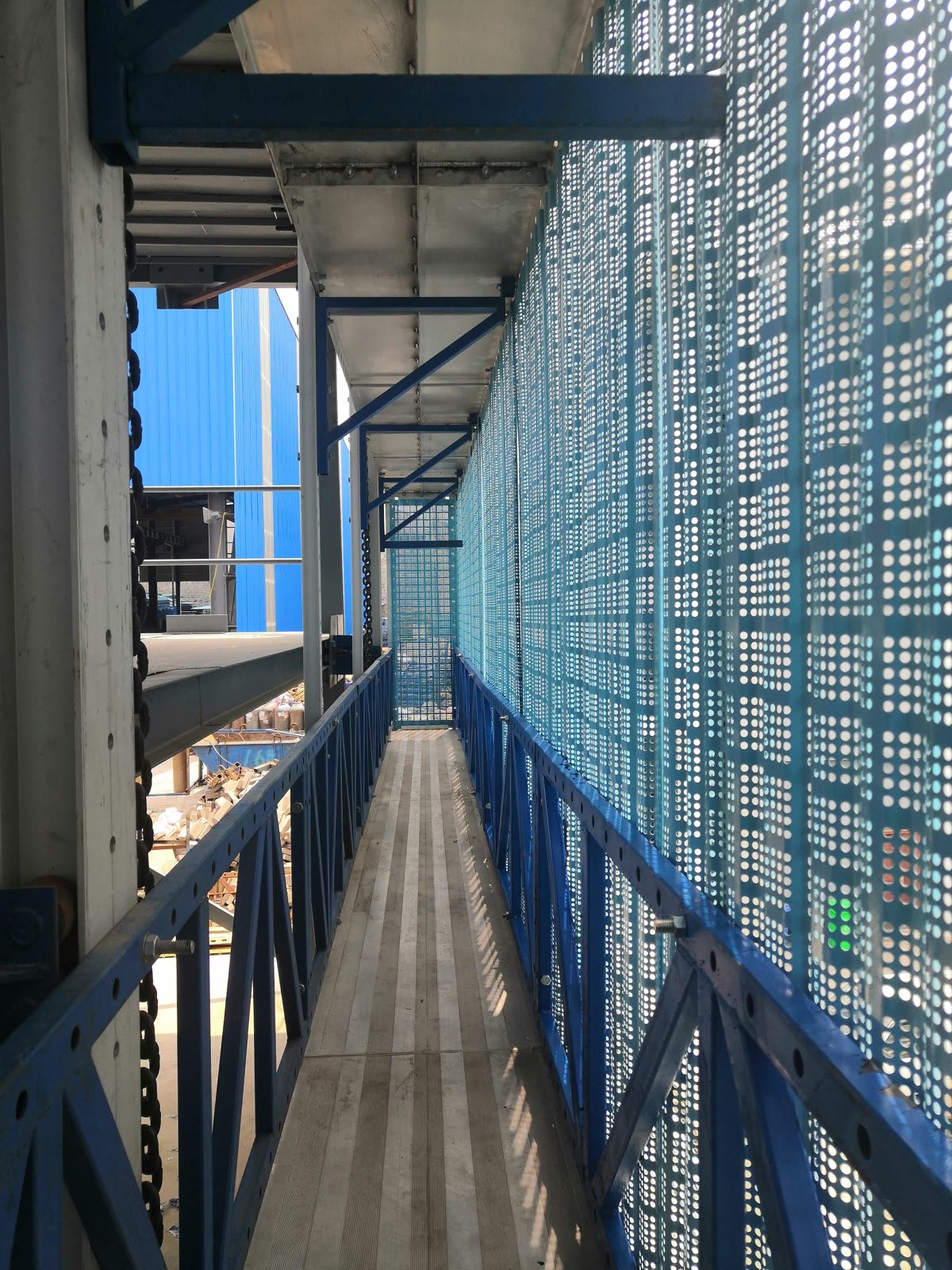 Inside a scaffold walkway: ribbed metal decking, blue side trusses with triangular braces and bolts, and a perforated outer screen.