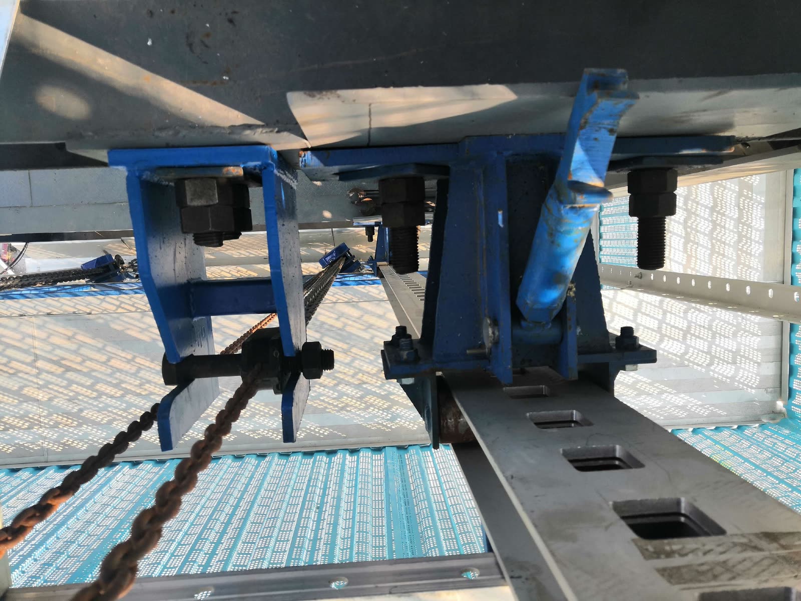 Close-up from below of a slotted rail, blue support brackets, large threaded fasteners and hoist chains.