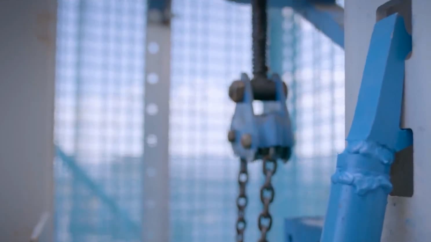 A hoist chain and hook hanging beside a blue support member with a forked top, inside the enclosure.