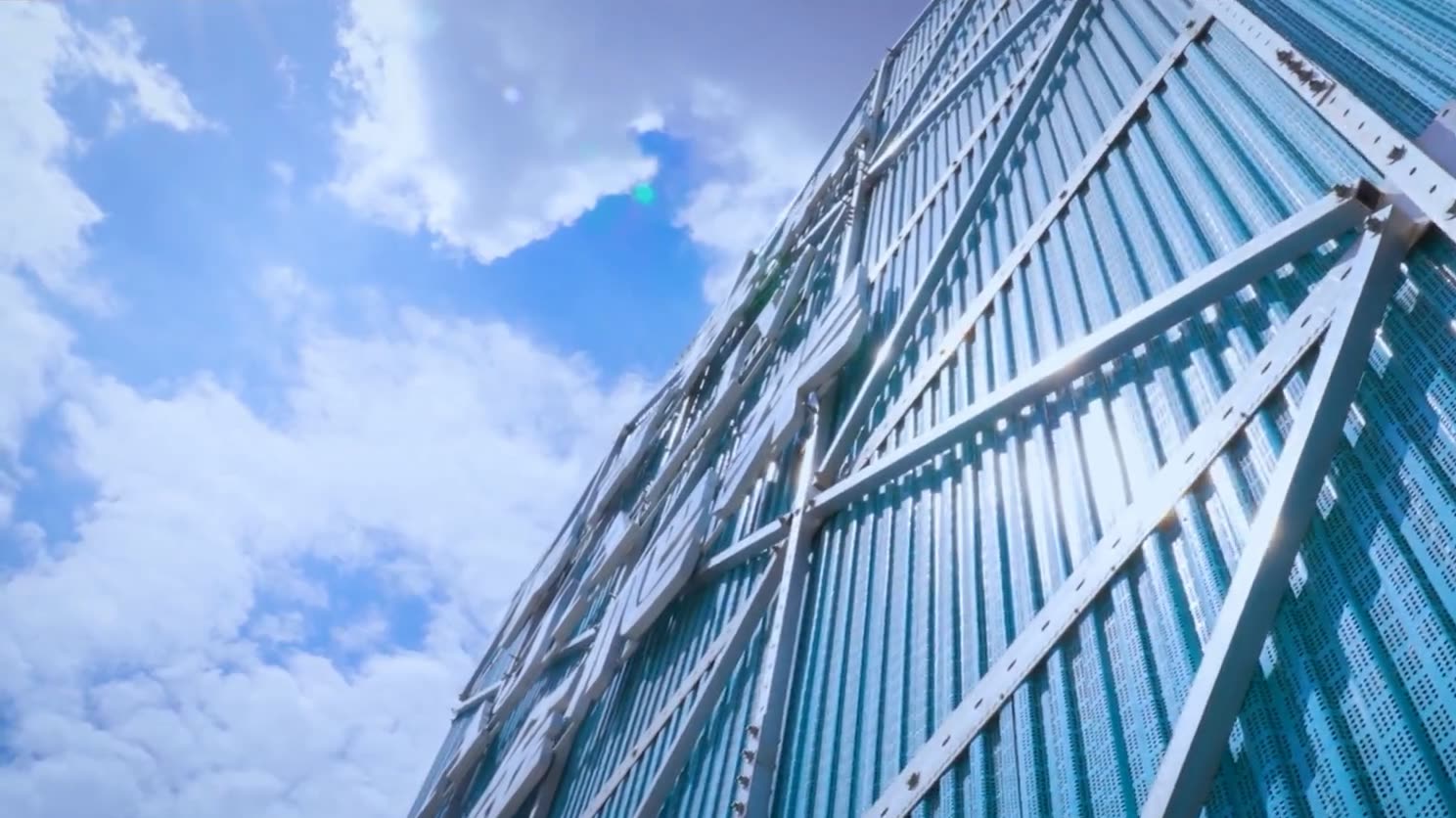 Looking up at a climbing-scaffold enclosure: silver frame members and diagonal braces over blue perforated mesh panels, against a blue sky.