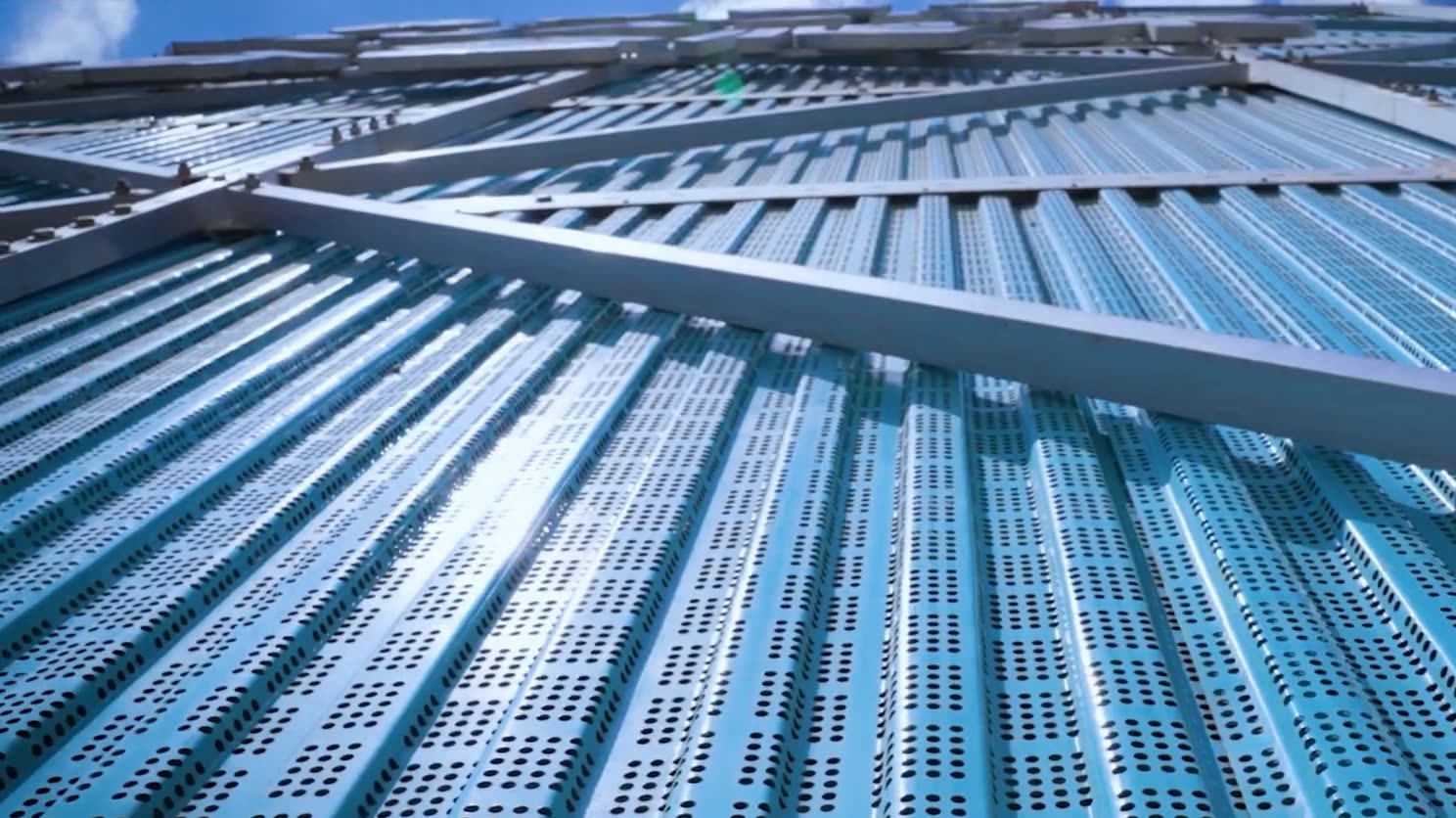 Close-up of blue perforated, corrugated mesh panels on the enclosure, with frame members across them and sunlight through the perforations.